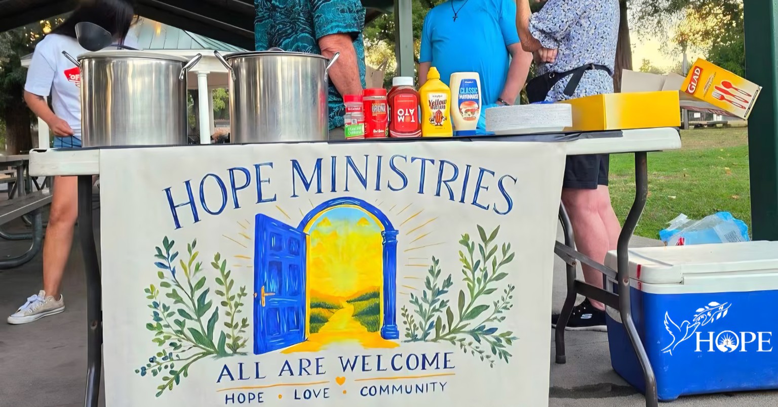Hope Ministries outreach table with volunteers and All Are Welcome banner